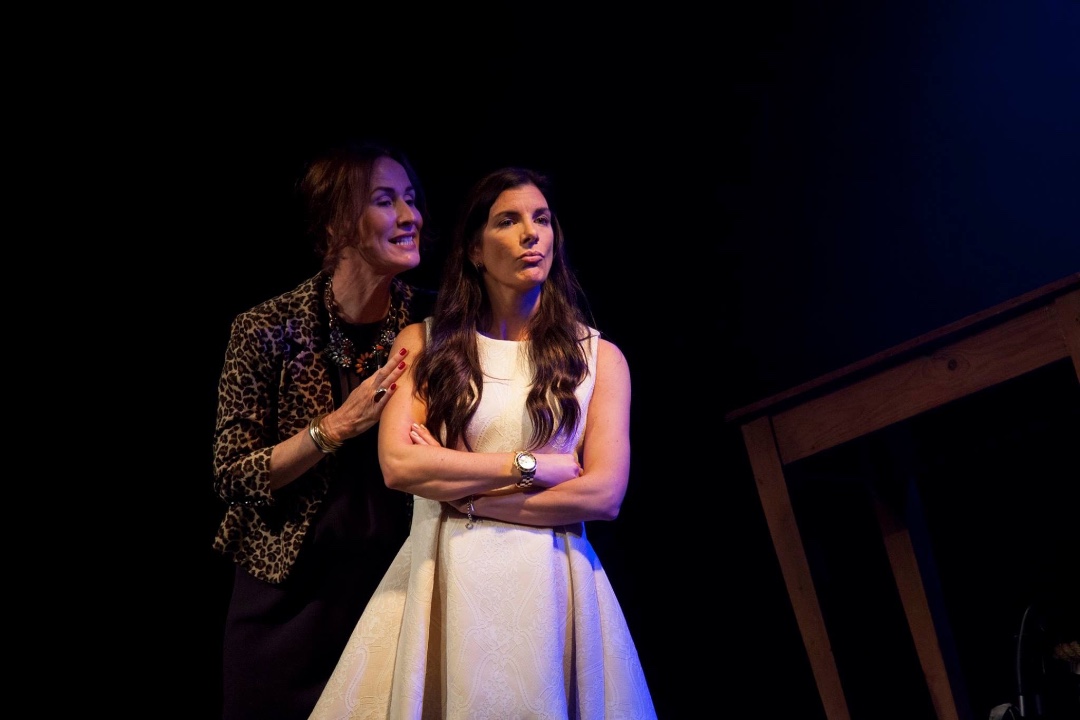 A woman in a leapord skin jacket stands closely behind another woman in a white strappy dress, with her right hand on the shoulder of the woman in white. The woman in white is stood with a defiant look on her face and her arms folded in front of her.