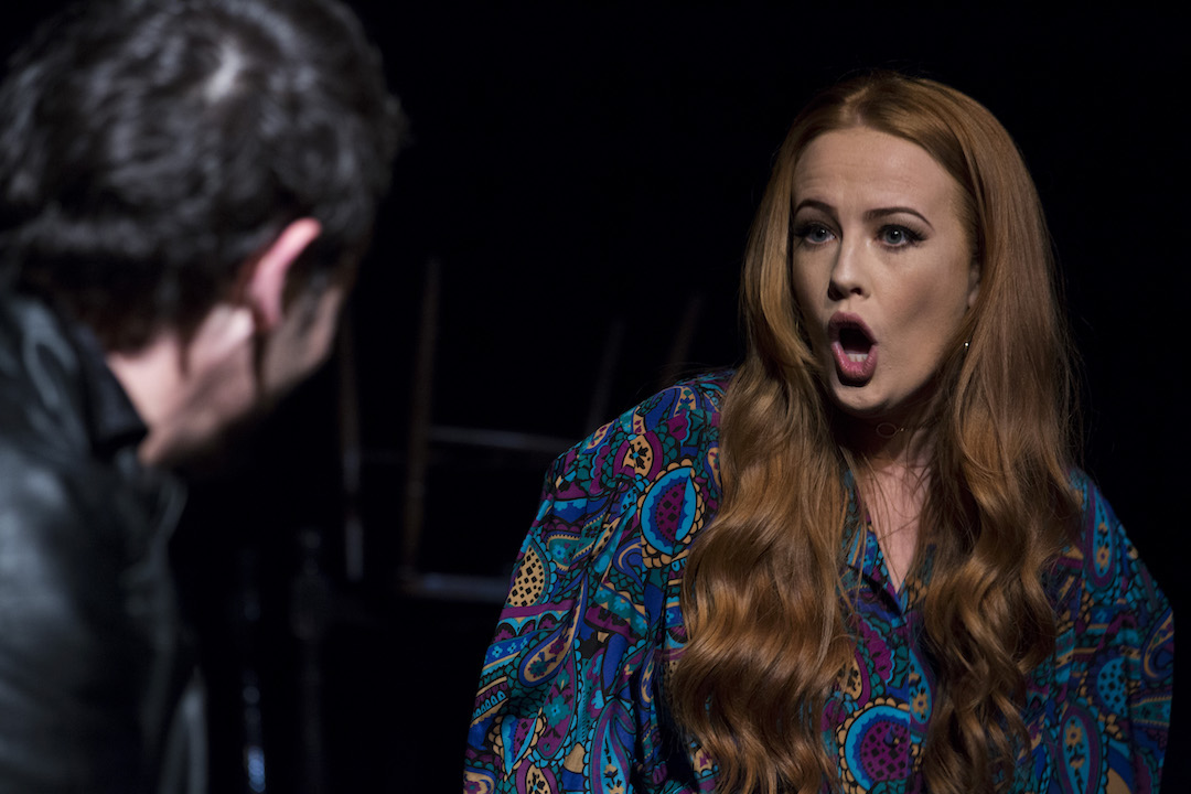 An out of focus man with black hair and a black leather jacket, is looking into the picture (we only see the back of his head). There is a red headed woman, her long hair flowing either side of her face to lay on her chest, looks back at the man, mouth wide open in an expression of shock or shouting.