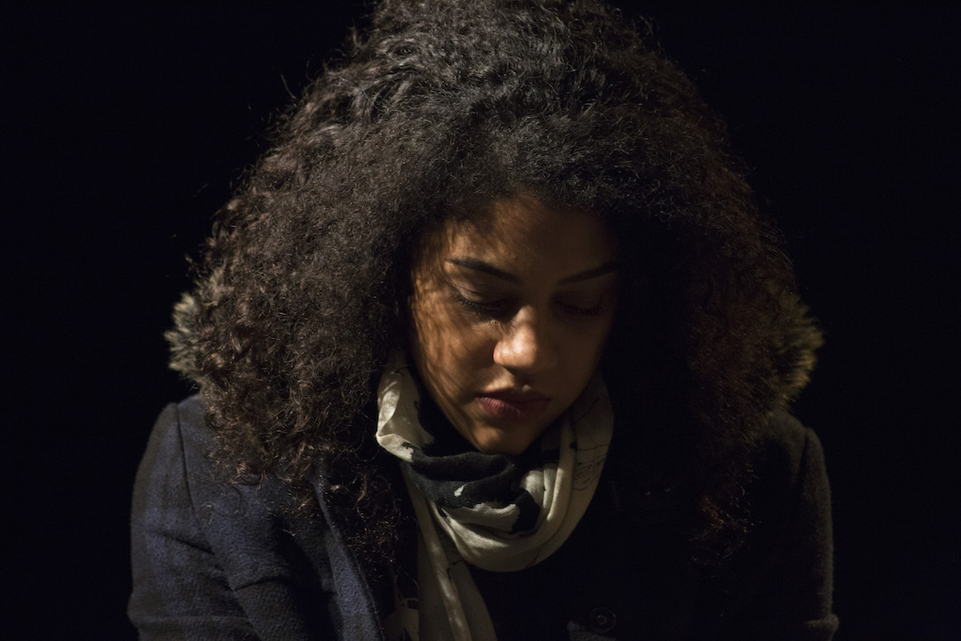 An upper torso and head shot of a young woman, looking down, with half of her obscured in shadows.