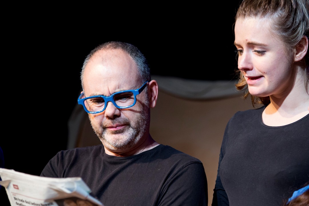 A blading man in a black t-shirt and blue rimmed glasses, looks at a news paper in his hands. Next to him is a taller youg woman wearing a long sleeved, tight-fitting, black t-shirt, looking at the news paper with a slightly confused look on her face.