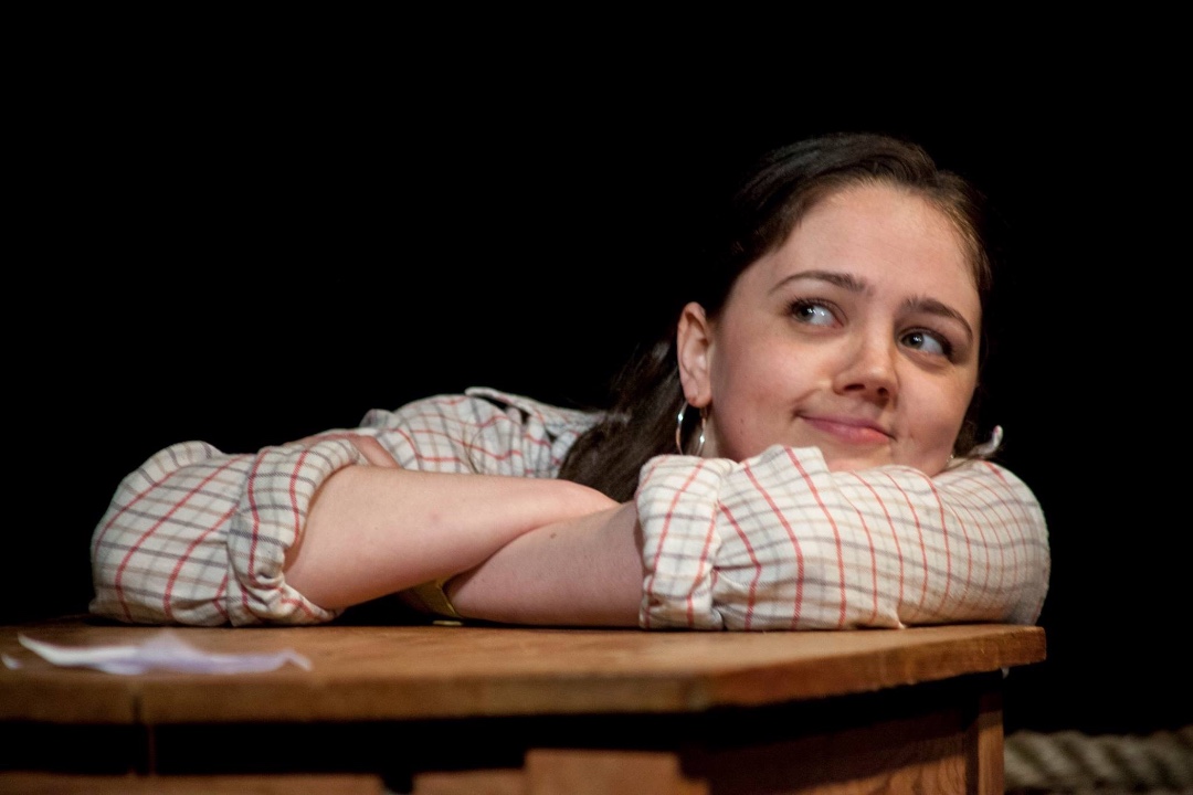 A young woman, with a cheeky smile on her face, is kneeling at a desk with her arms cross on it, while she looks up at someone out of shot.