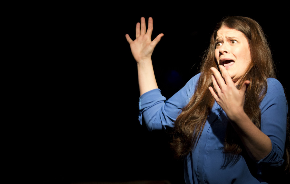 A woman in a blue shirt, with the sleeves rolled up to her elbows. He right hand is up in the air palm out, her left hand is in front of her face palm in. She has an expression of surprise or shock on her face.