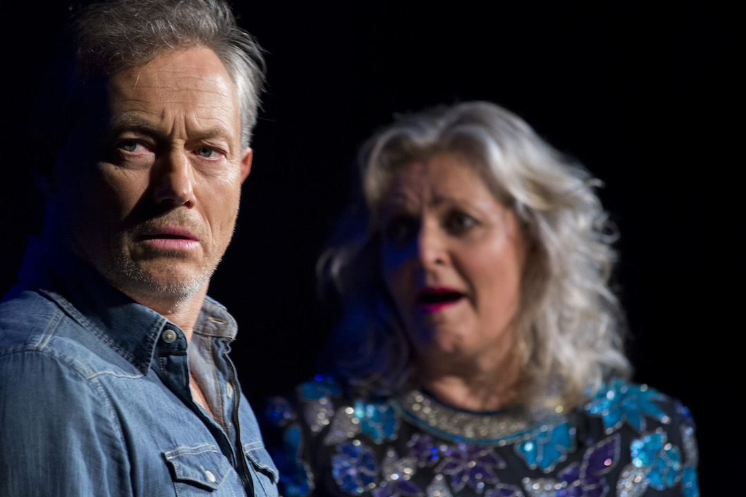 A man with grey hair and grey stubble, wearing a demin shirt, is the main focus of the image. He is in a side profile, looking a the camera. Out of focus behind him, in front profile, is a woman with shoulder length grey hair. She is looking at the man with an expression of shock on her face.