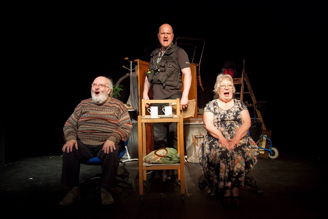 An elderly couple sit either side of a small table with cups of tea on it, shouting out something loud. A Policeman stands between them, behind the table, also shouting something.